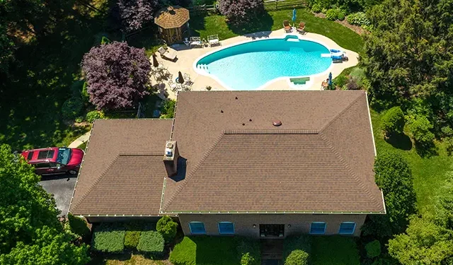 Aerial view of a single-story house with a brown roof, green lawn, and a curved blue pool in the backyard surrounded by trees and deck area.