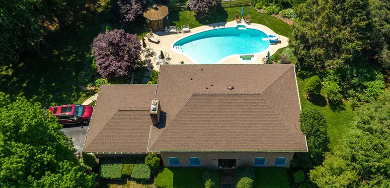 Aerial view of a single-story house with a brown roof, green lawn, and a curved blue pool in the backyard surrounded by trees and deck area.