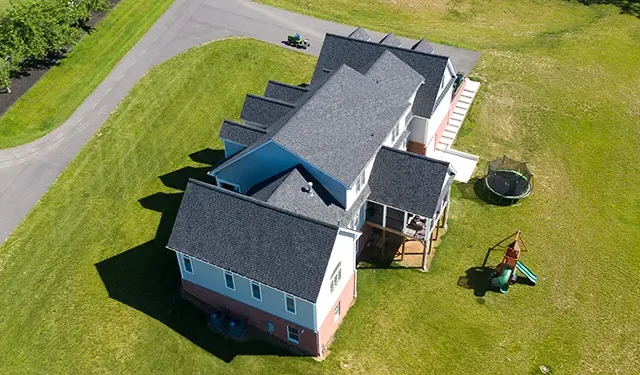 Aerial view of a large multi-wing house with gray shingle roof, light blue and brick red exterior, surrounded by green lawn and curved driveway nearby. Several sunlit shadows on the yard.