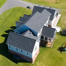 Aerial view of a large multi-wing house with gray shingle roof, light blue and brick red exterior, surrounded by green lawn and curved driveway nearby. Several sunlit shadows on the yard.