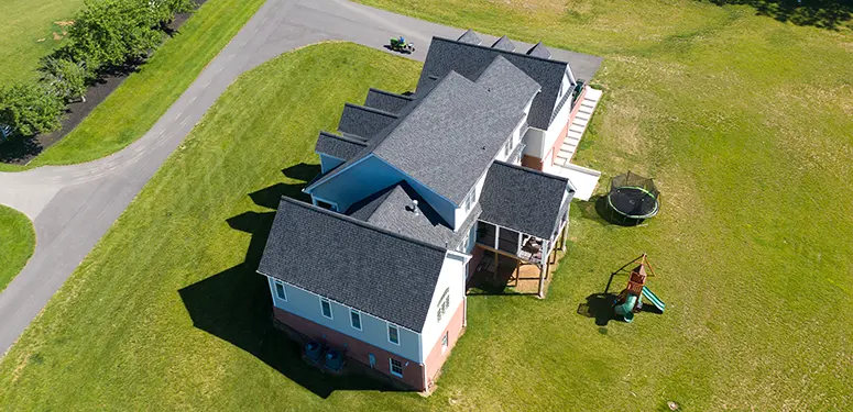 Aerial view of a large multi-wing house with gray shingle roof, light blue and brick red exterior, surrounded by green lawn and curved driveway nearby. Several sunlit shadows on the yard.
