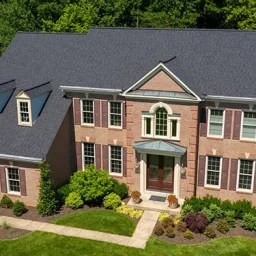 Aerial view of a two-story brick home with dormer windows, a centered front door, and a manicured lawn with shrubs.