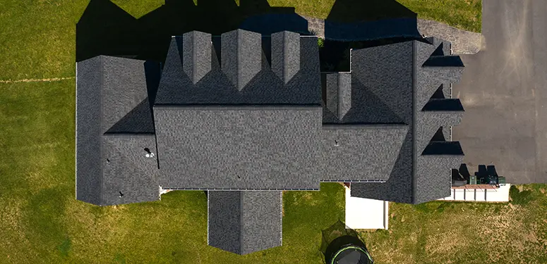 Aerial top-down view of a large gray-shingled house with multiple gabled roofs, surrounding lawn, and a paved driveway on the right.
