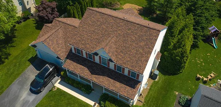 Aerial view of a two-story house with a brown shingle roof, white siding, a blue car in the driveway, and a green lawn with trees nearby.