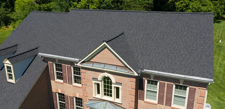 Aerial view of a two-story brick house with dark shingled roof, white trim, and brown shutters surrounded by green lawn.
