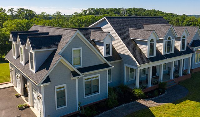 Two-story suburban house with multiple gabled roofs, light blue siding, front porch, and surrounding green lawn.