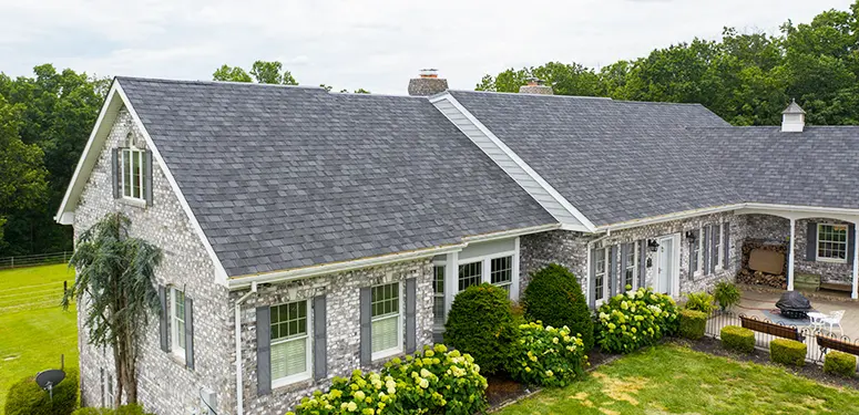 Stone ranch-style house with a gray shingle roof, brick-like siding, and manicured shrubbery in a green yard.