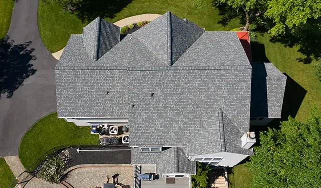 Aerial top-down view of a gray shingle-roofed house with surrounding lawn, trees, and curved driveway nearby.