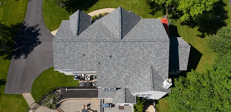 Aerial top-down view of a gray shingle-roofed house with surrounding lawn, trees, and curved driveway nearby.