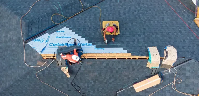 Overhead view of two workers installing roofing on a sloped roof, laying blue underlayment and dark shingles with tools nearby