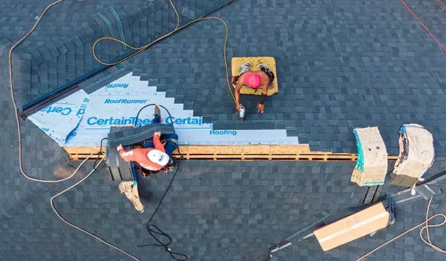Overhead view of two workers installing roofing on a sloped roof, laying blue underlayment and dark shingles with tools nearby