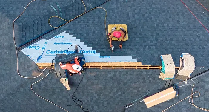 Overhead view of two workers installing roofing on a sloped roof, laying blue underlayment and dark shingles with tools nearby