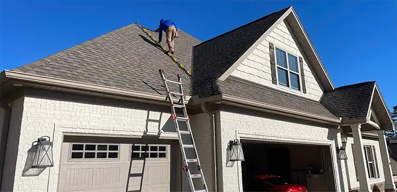 Worker on the sloped roof performing maintenance on shingles; a ladder leans against a white brick house with an open garage and bright blue sky above.