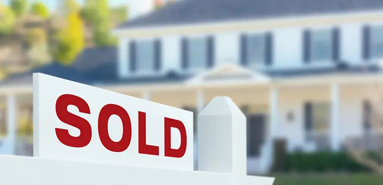 “Sold” sign in front of a white picket fence with a blurred suburban house in the background, indicating a home has been sold.