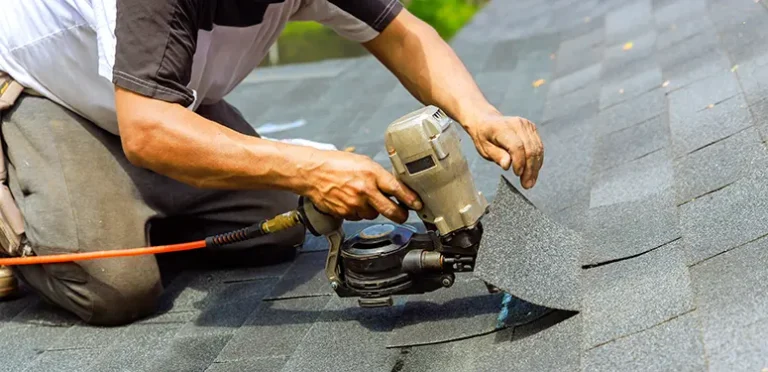 Roofer kneeling on asphalt shingles, operating a pneumatic nail gun to install or repair a roof.