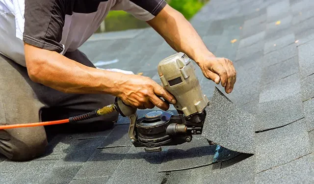 Roofer kneeling on asphalt shingles, operating a pneumatic nail gun to install or repair a roof.