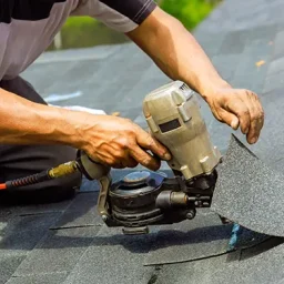 Roofer kneeling on asphalt shingles, operating a pneumatic nail gun to install or repair a roof.