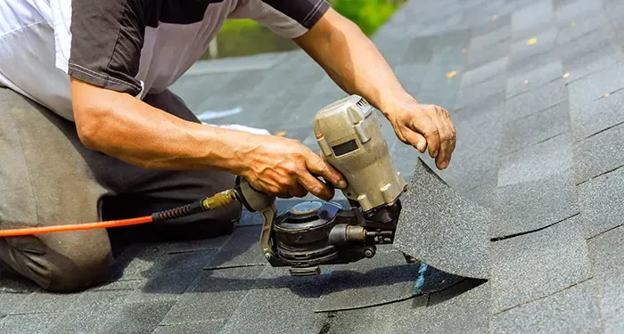 Roofer kneeling on asphalt shingles, operating a pneumatic nail gun to install or repair a roof.