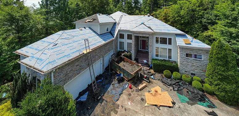 A two-story house under renovation with a partially completed roof and construction debris in the driveway