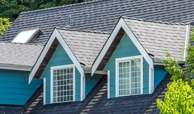 Blue house roof with two white-trimmed dormer windows and a dark shingled roof, surrounded by green trees.