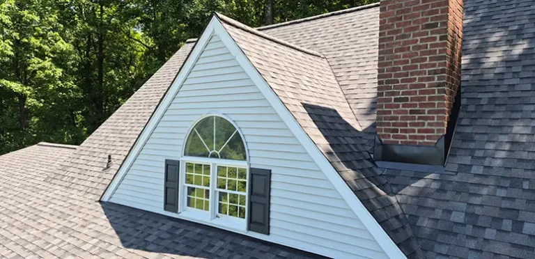 A dormer window with an arched top and open shutters on a gray shingled roof, surrounded by trees, with a brick chimney to the side.