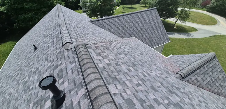 Aerial view of a gray shingled roof with curved ridge vents and a vent cap, surrounding lawns and a curved driveway nearby