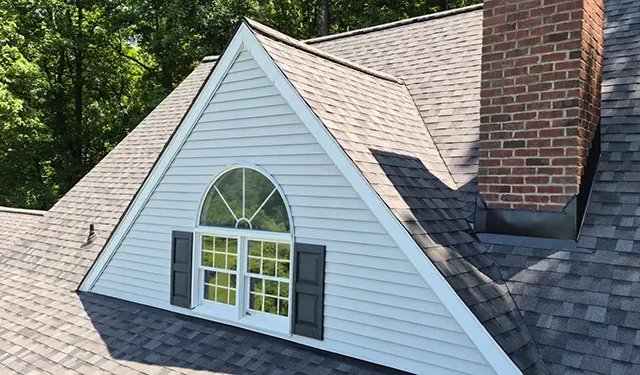 A dormer window with an arched top and open shutters on a gray shingled roof, surrounded by trees, with a brick chimney to the side.