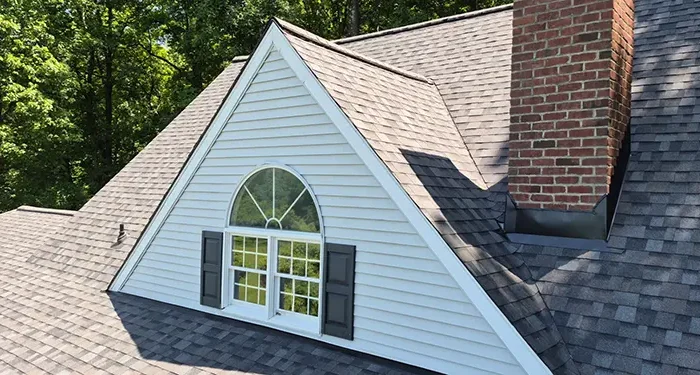 A dormer window with an arched top and open shutters on a gray shingled roof, surrounded by trees, with a brick chimney to the side.