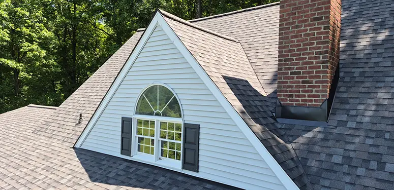 A dormer window with an arched top and open shutters on a gray shingled roof, surrounded by trees, with a brick chimney to the side.