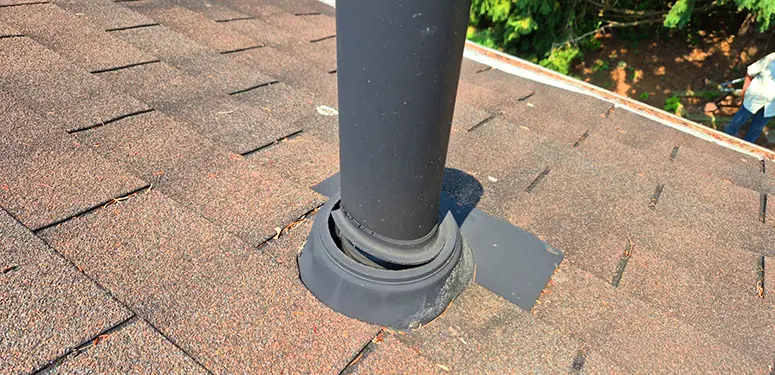 Black vent pipe protrudes from a brown shingled roof, surrounded by circular flashing; greenery visible in the background.