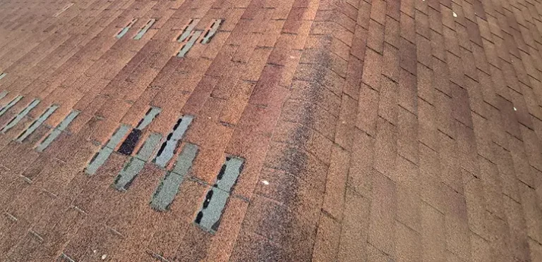 Close-up of a brown asphalt roof showing several cracked and missing shingles with exposed underlying material (teal/blue patches).