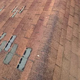 Close-up of a brown asphalt roof showing several cracked and missing shingles with exposed underlying material (teal/blue patches).
