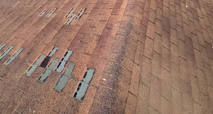 Close-up of a brown asphalt roof showing several cracked and missing shingles with exposed underlying material (teal/blue patches).